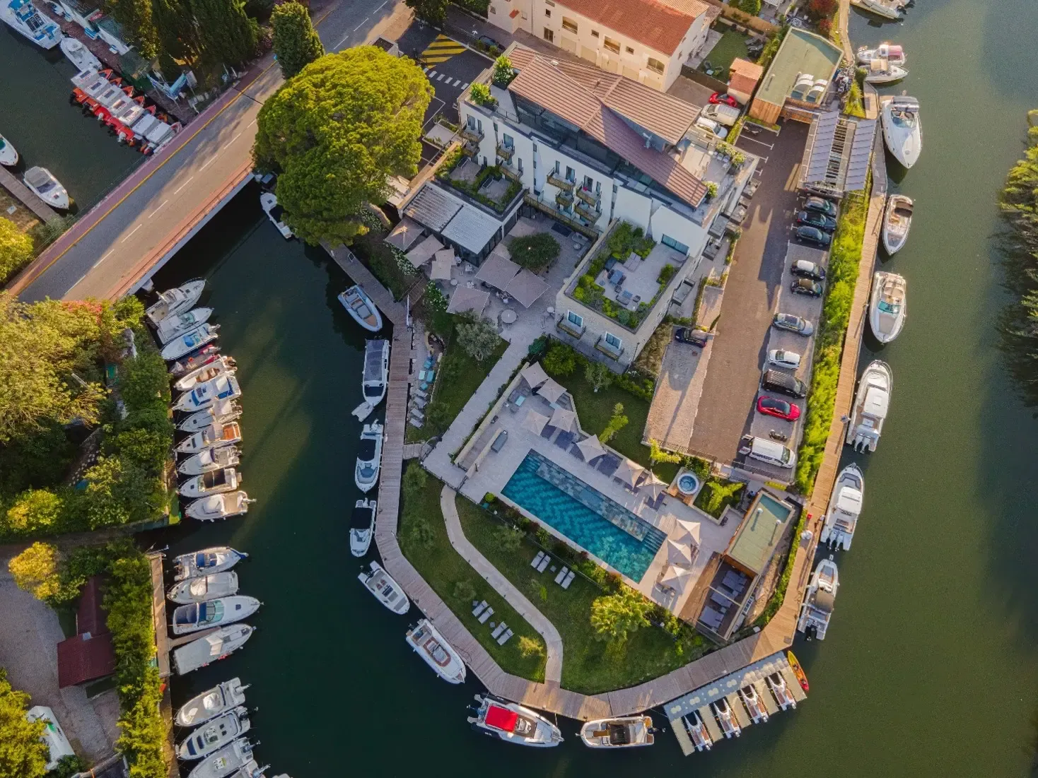 Aerial view of a riverside residential property with a swimming pool, moored boats, and parked cars