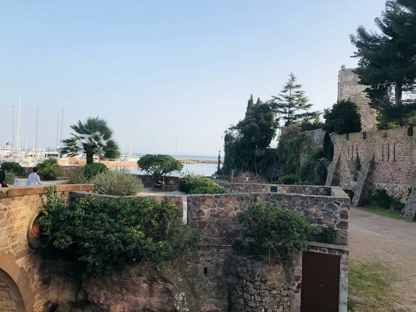 View of a stone fortress with ancient walls, plants, and trees, overlooking a harbor with boats