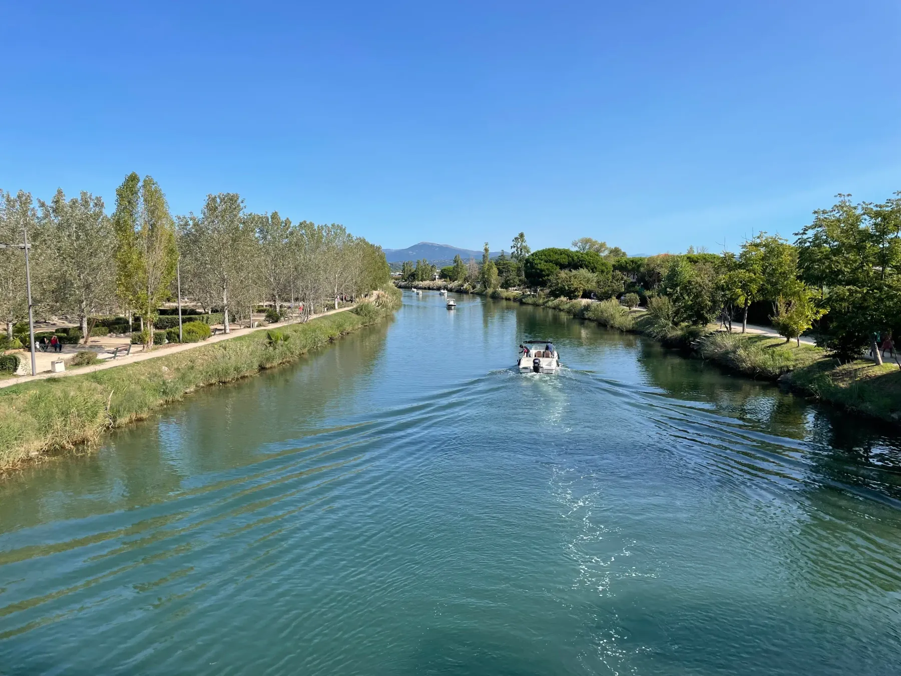 A boat sails on a river lined with trees and walking paths under a blue sky.