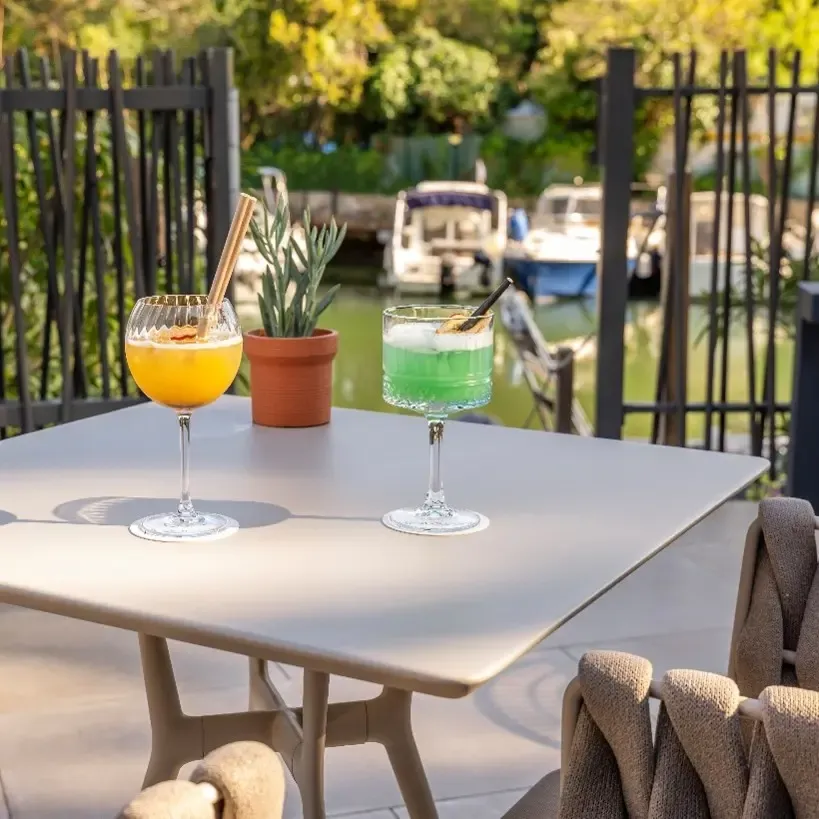Two colorful cocktail glasses on a terrace table near a canal with boats, surrounded by plants and chairs.