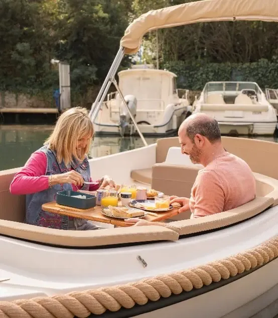 A couple is having a meal on a boat moored in a canal, with dishes and drinks arranged on a table between them.