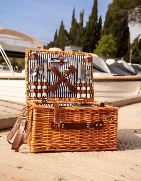 Two wicker baskets with leather straps are placed on a dock near moored boats.