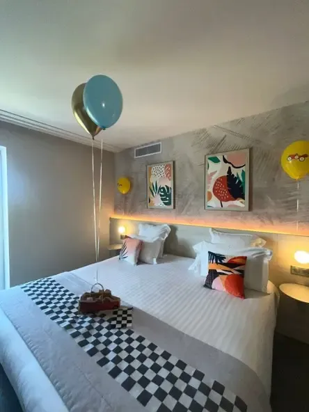 Room with a double bed covered by a black and white checkered blanket, two colorful paintings on the wall, balloons and a box of chocolates on the bed