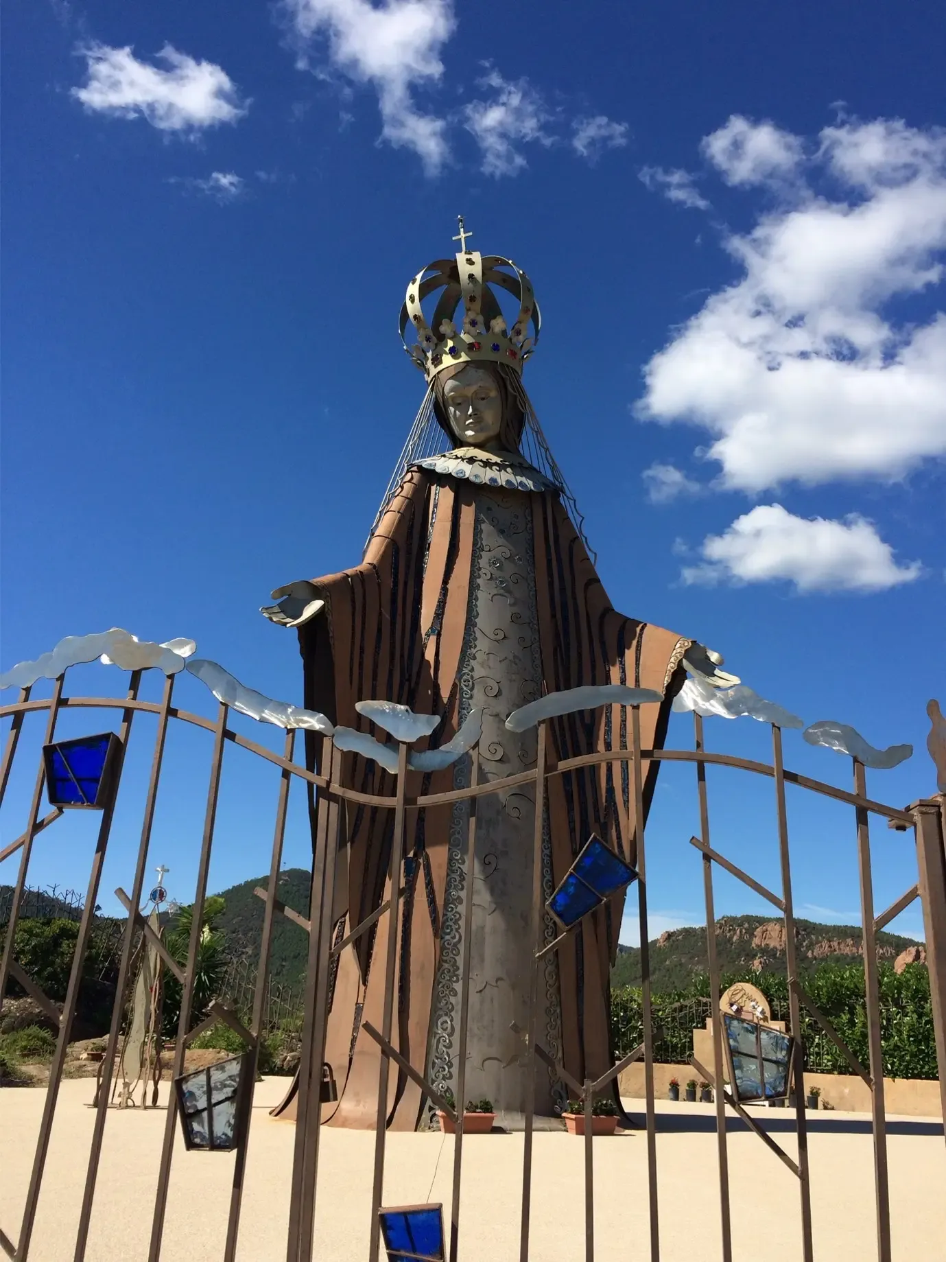 Metal statue of a crowned religious figure, surrounded by an iron structure with colored glass elements, under a blue sky with clouds