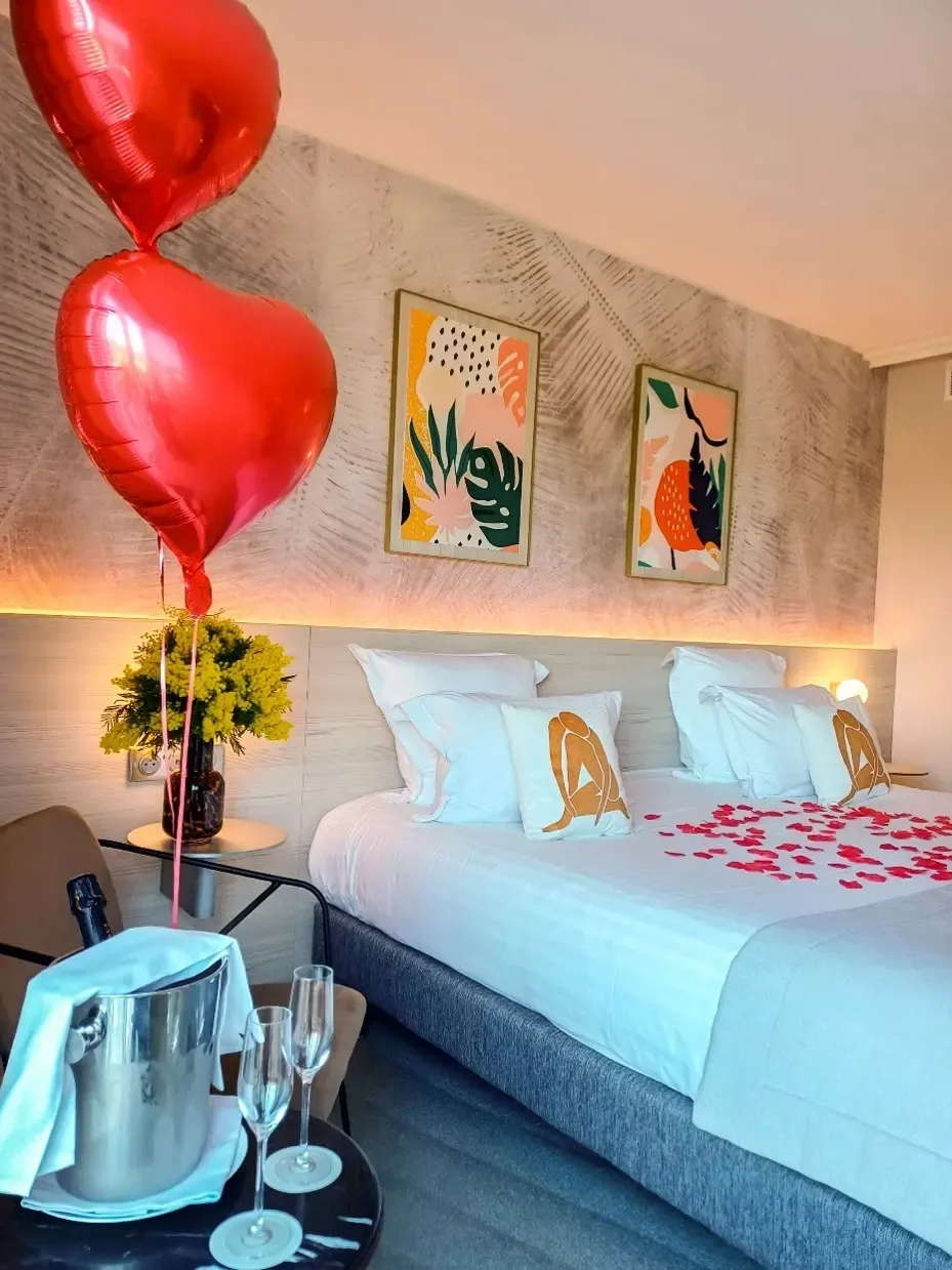 Room with a bed covered in red petals, two heart-shaped balloons, a vase with flowers, a bottle of champagne in an ice bucket, and two champagne flutes on a table.