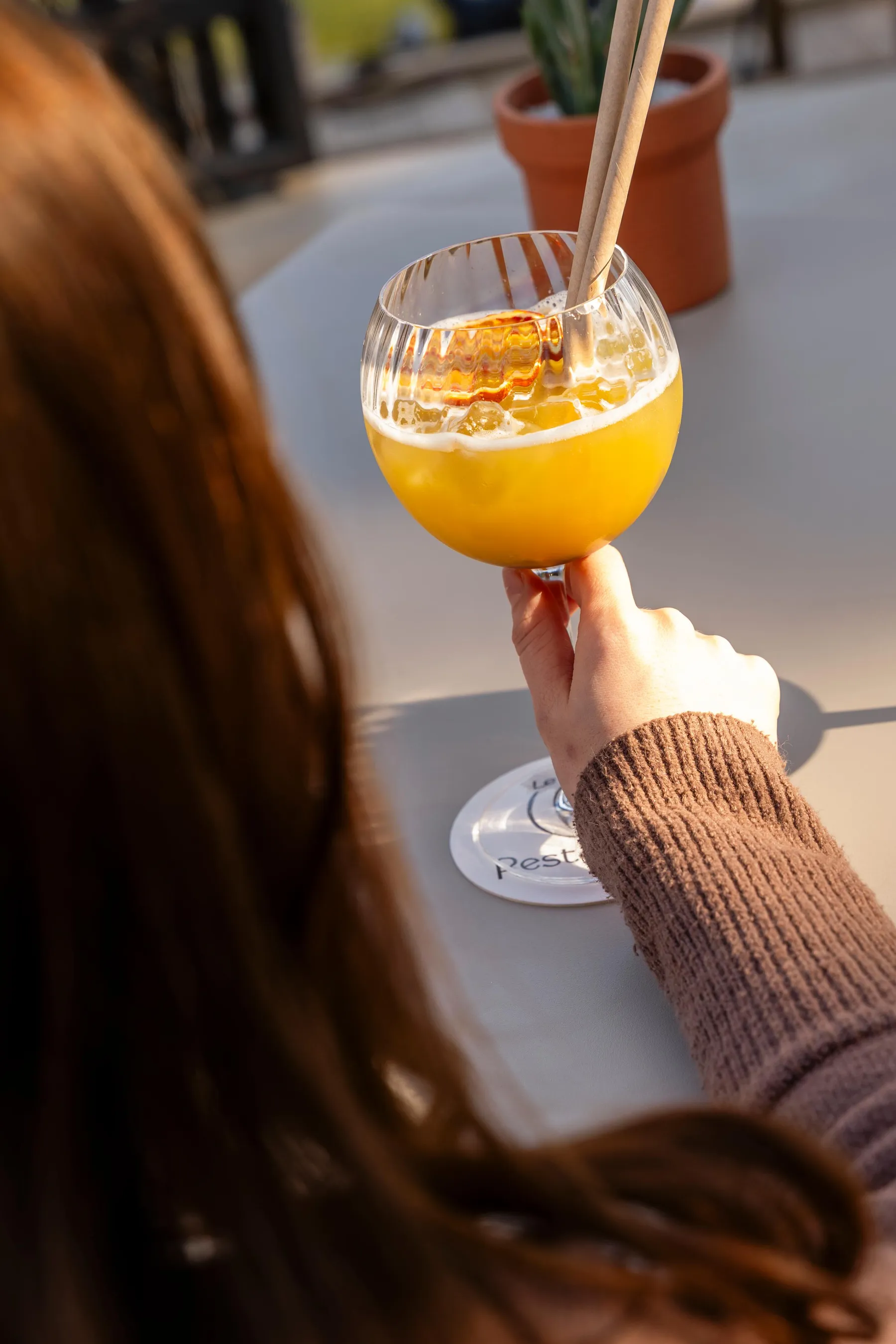 A hand holding a glass containing a yellow drink with ice cubes and a straw, placed on an outdoor table with flower pots in the background.