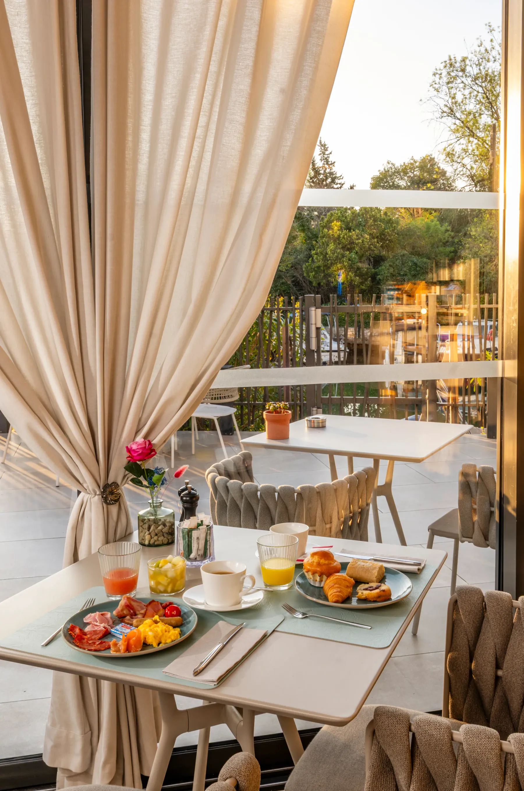 Table set for breakfast with charcuterie platters, croissants, fruit, drinks, and a rose in a vase, viewed from inside a restaurant with curtains and an outdoor terrace visible through a glass door.