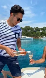 A man in a striped T-shirt pours white wine into a glass on a motorboat by the water.