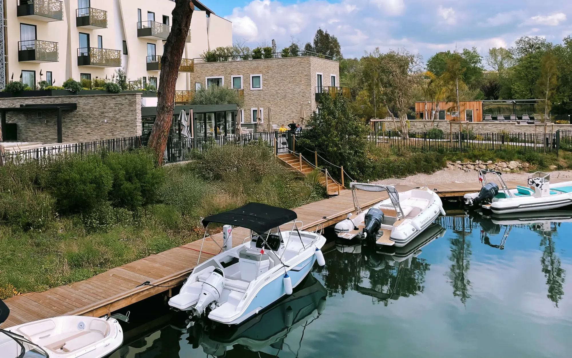 Wooden quays with several small motorboats moored in front of modern residential buildings surrounded by vegetation
