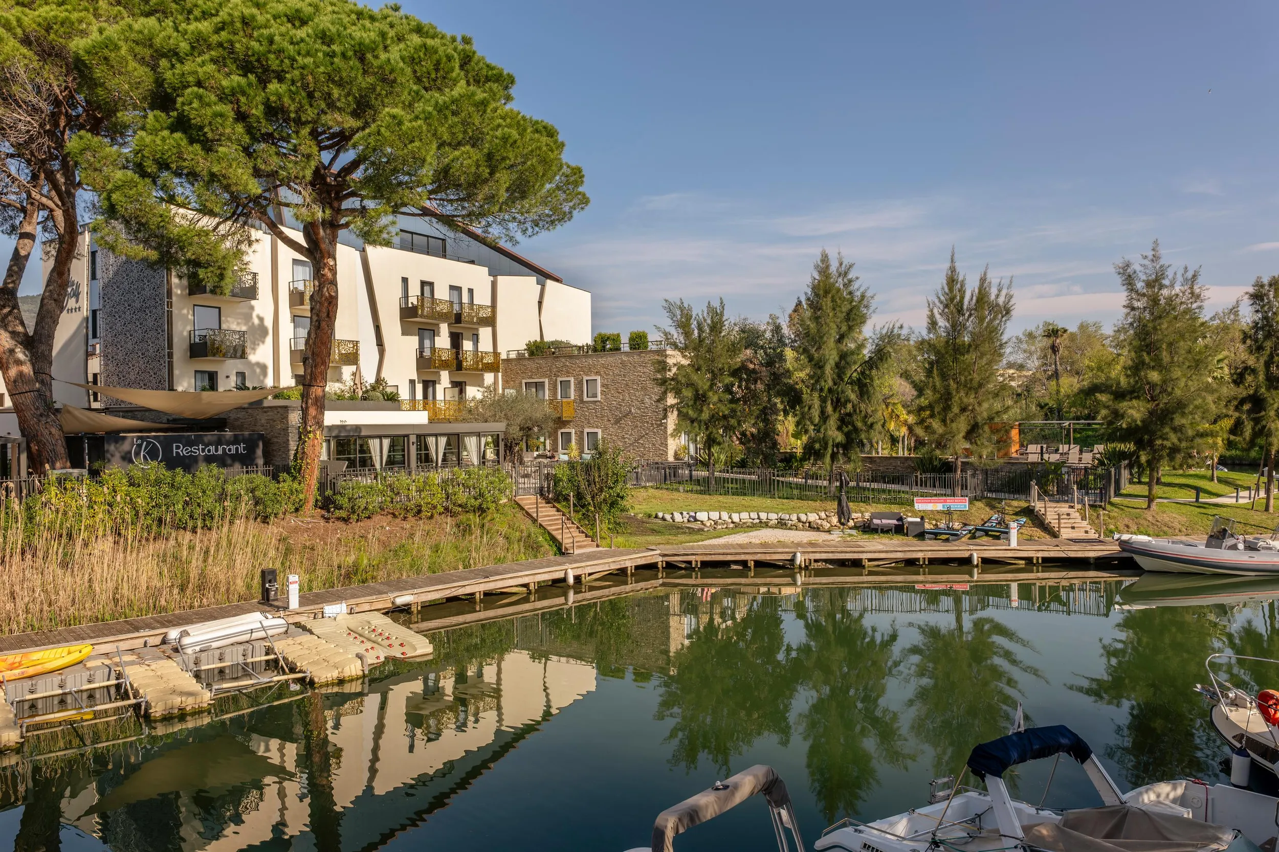 A building with a restaurant and balconies overlooks a tree-lined canal with moored boats.