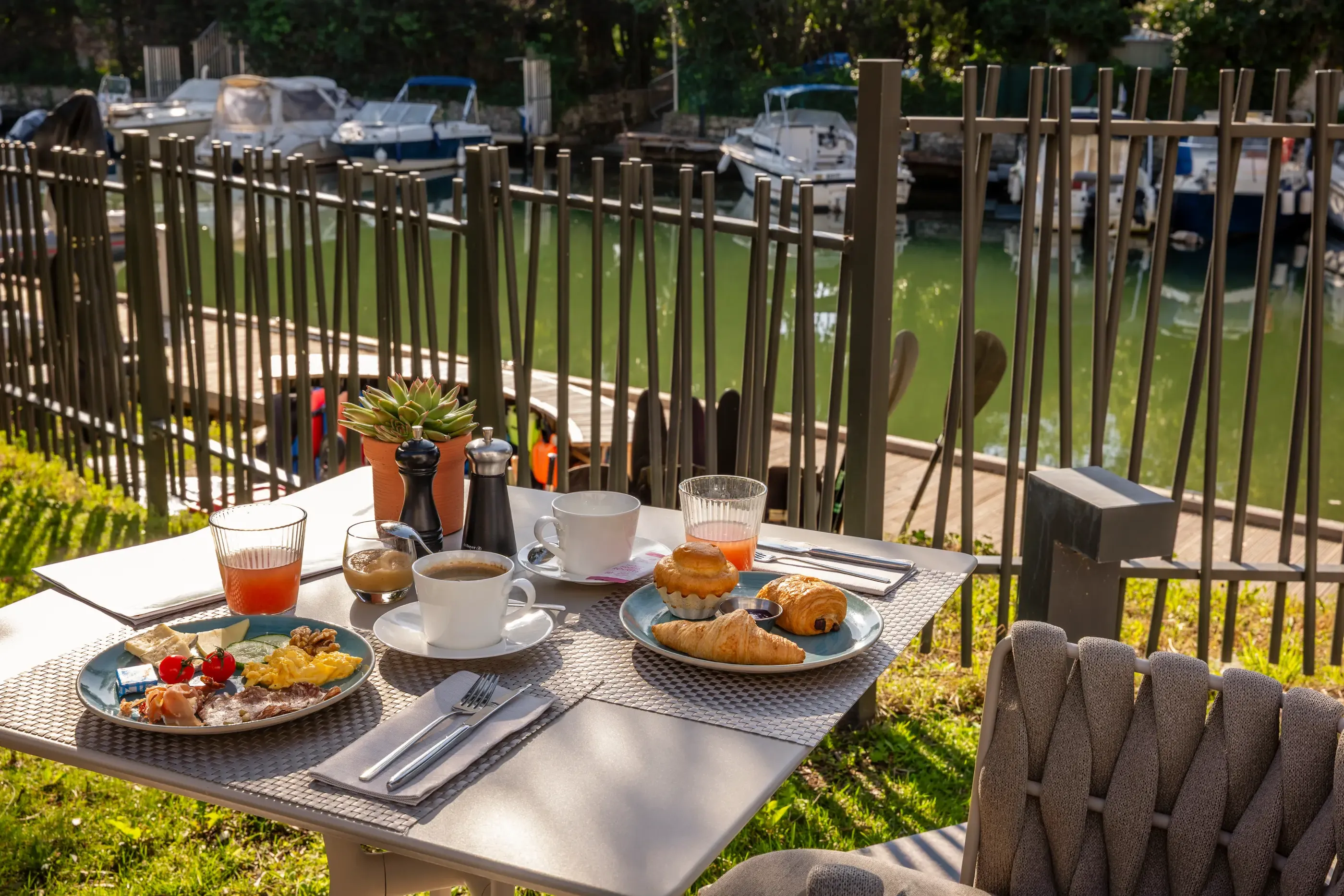 Outdoor table with breakfast consisting of croissants, bread, jam, fruit, and drinks, view of a canal with boats