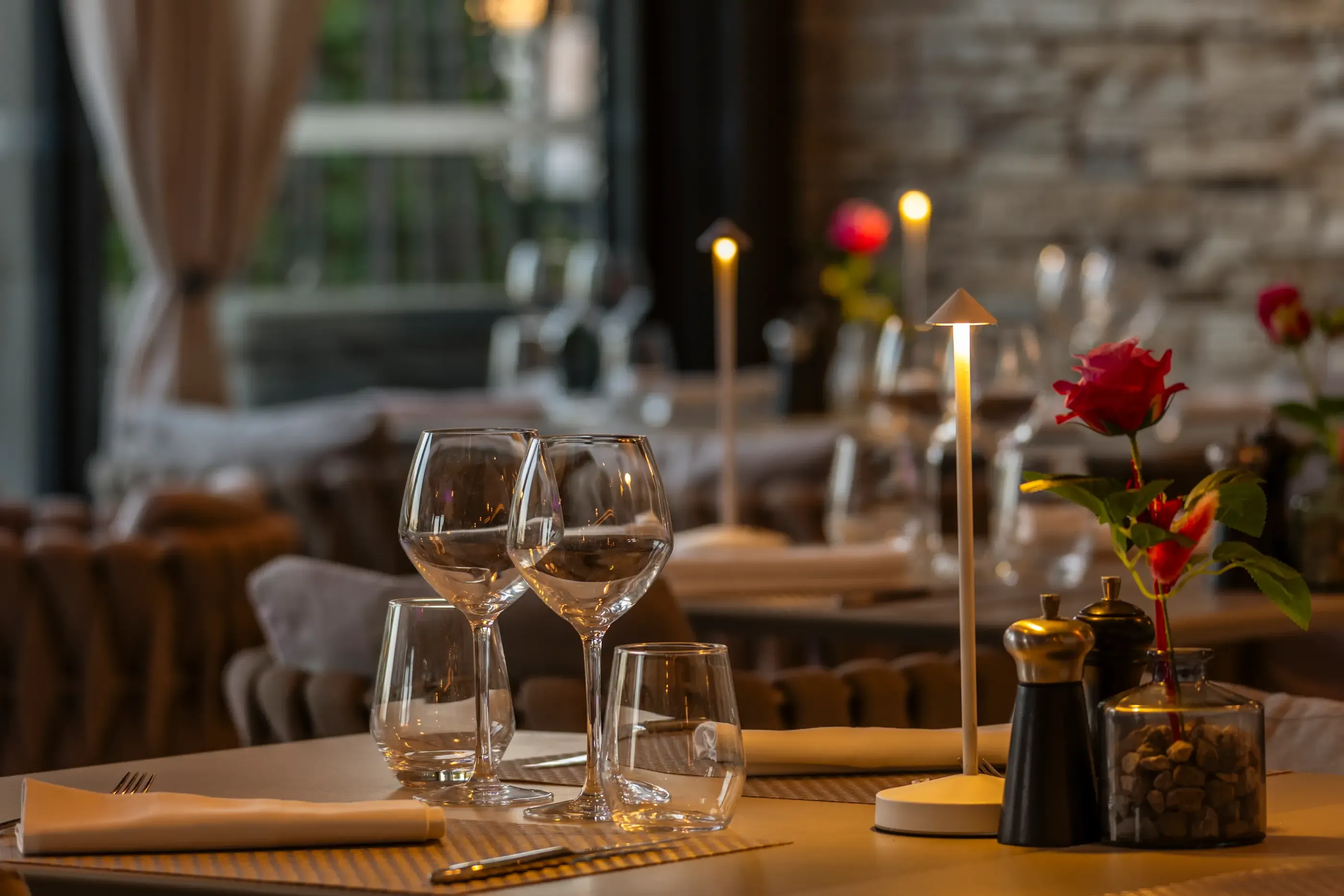 Table set with glasses, tablecloth, red rose, and candles in a dimly lit restaurant.