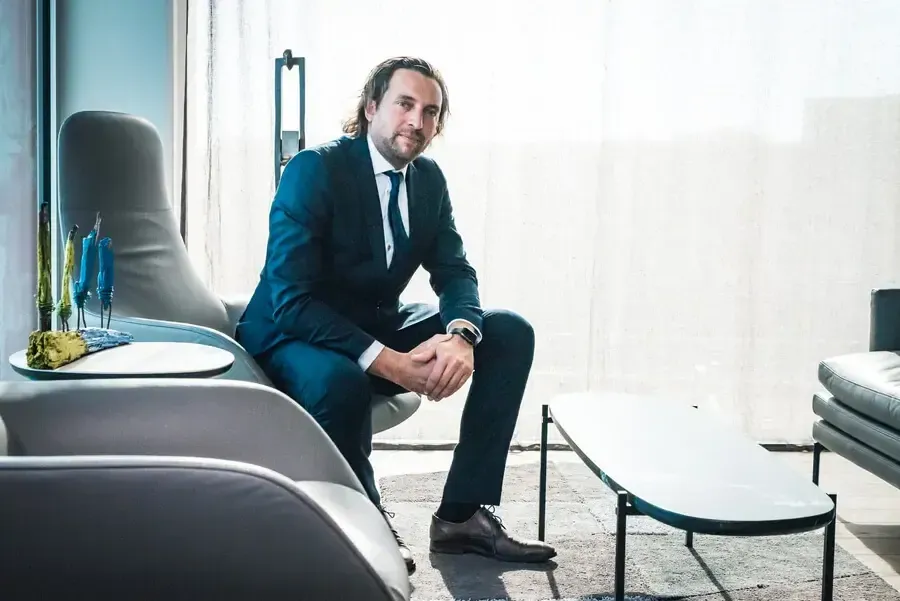 A man in a blue suit is sitting on a gray sofa in a modern space with a coffee table and decorative plants.