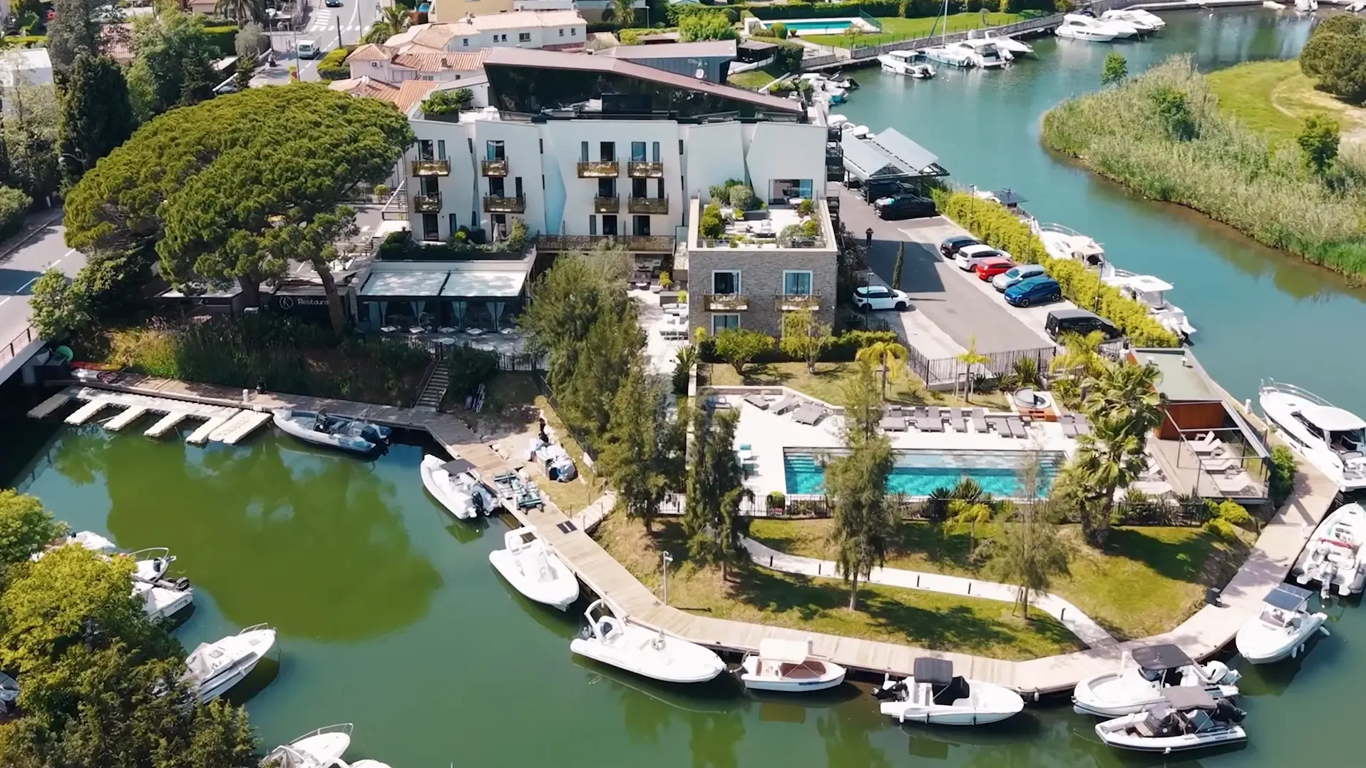 Aerial view of a hotel complex with a pool and marina, surrounded by water and moored boats.