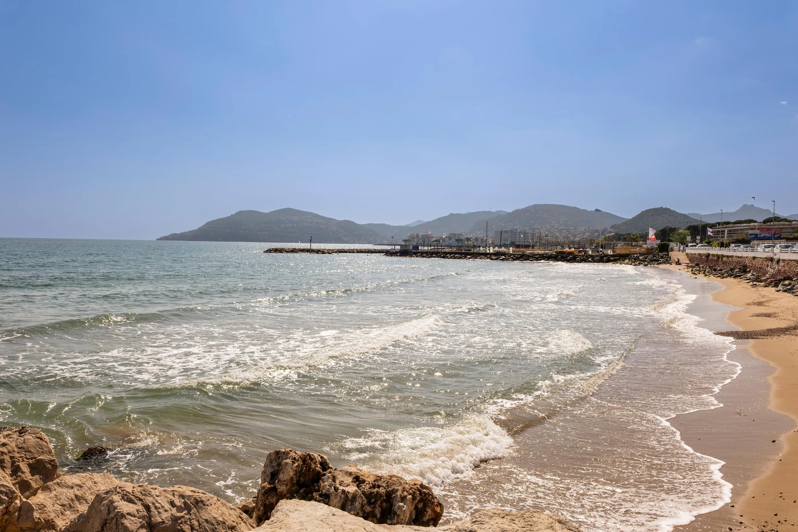 Small wave on a sandy beach with rocks, bordered by a calm sea and hills in the background.