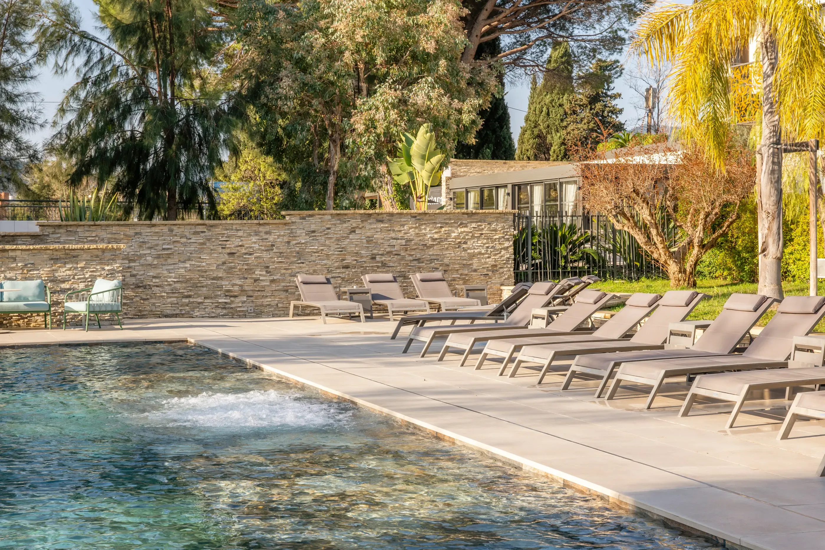 A pool with sun loungers lined up and a stone wall in the background, surrounded by trees and vegetation.