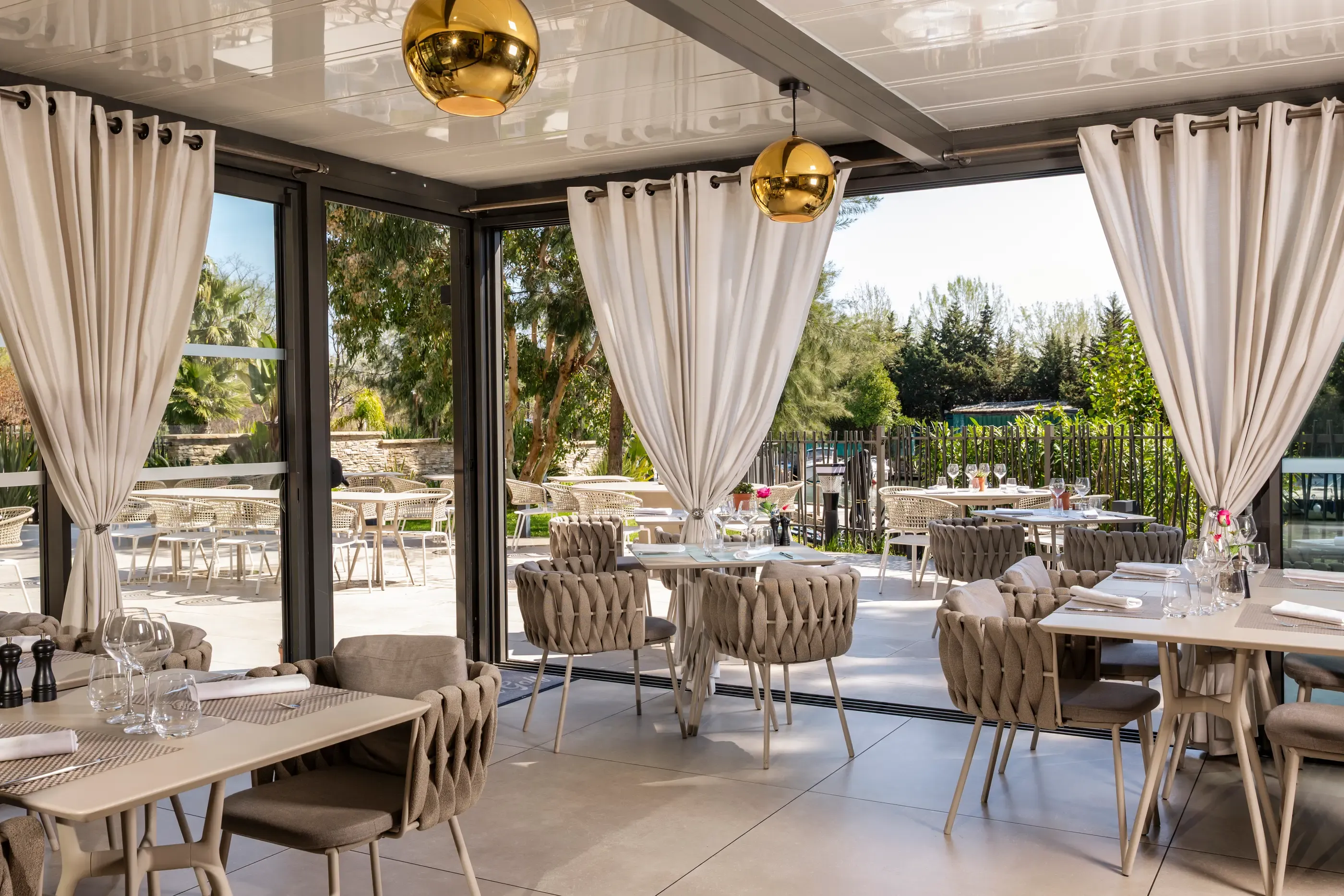 Restaurant room with tables and chairs arranged, large glass windows open onto an outdoor terrace, decorated with plants and spherical light fixtures.