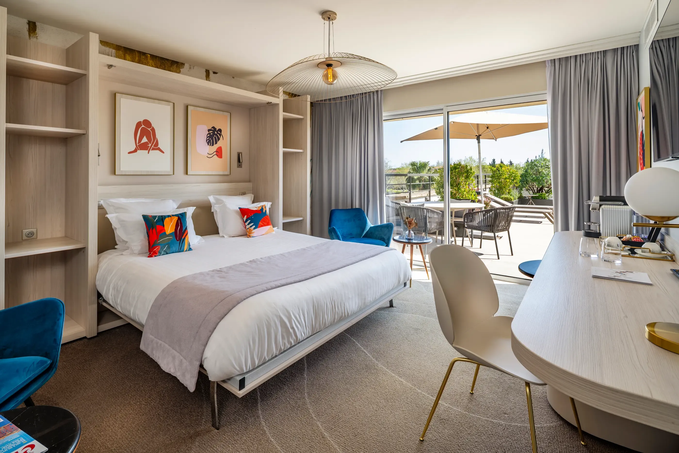 Modern hotel room with a double bed, a work desk, a blue armchair, and a view of an outdoor terrace with tables and chairs.