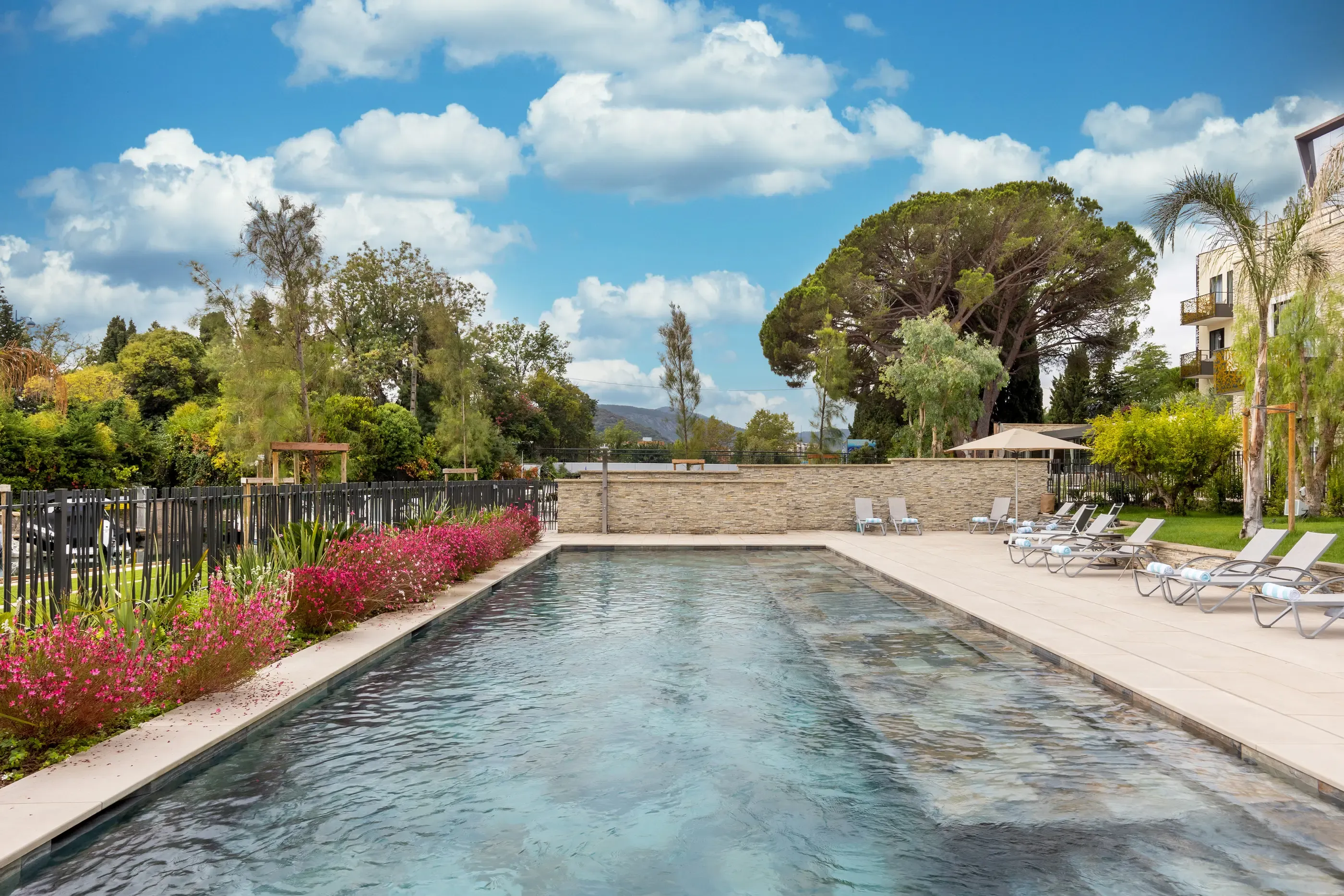 Rectangular swimming pool surrounded by lounge chairs and vegetation, with a partly cloudy sky in the background