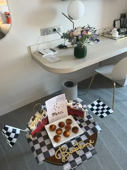 A tray with cakes and popcorn is placed on a black-and-white checkered blanket in front of a desk with a vase of flowers.