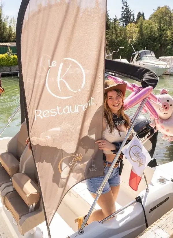 A woman smiles on a boat with a sign "Le K Restaurant" and a flag in the background.