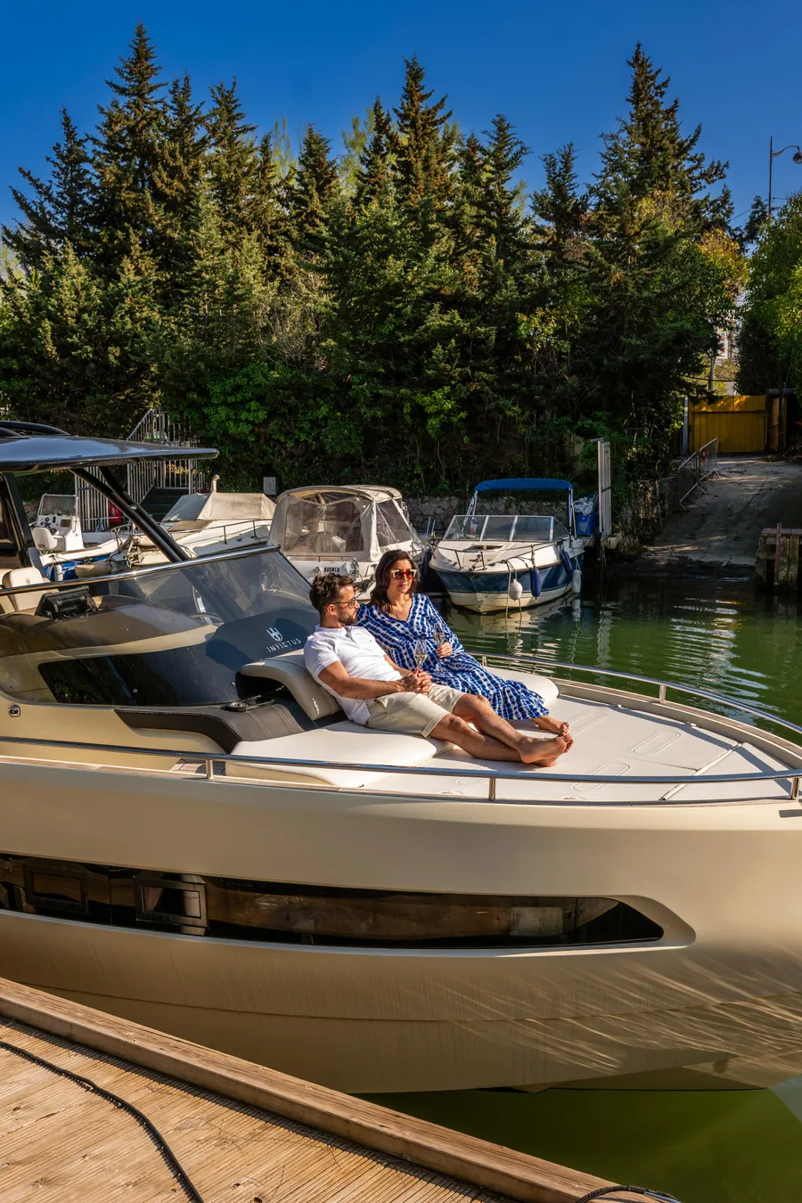 Two people sitting on a motorboat moored in a canal lined with trees and other boats.