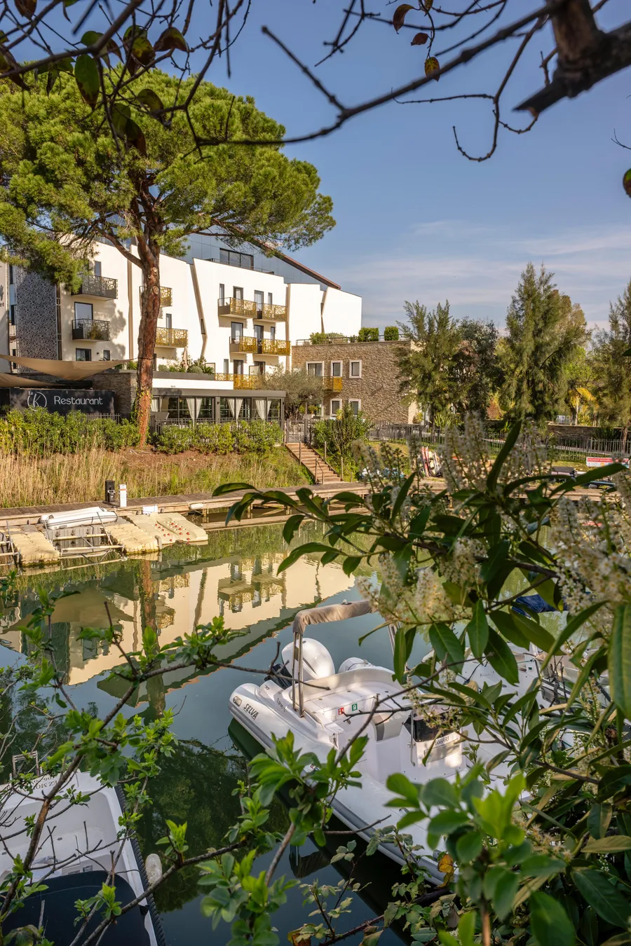 Boats moored on a canal lined with vegetation, with modern buildings in the background.