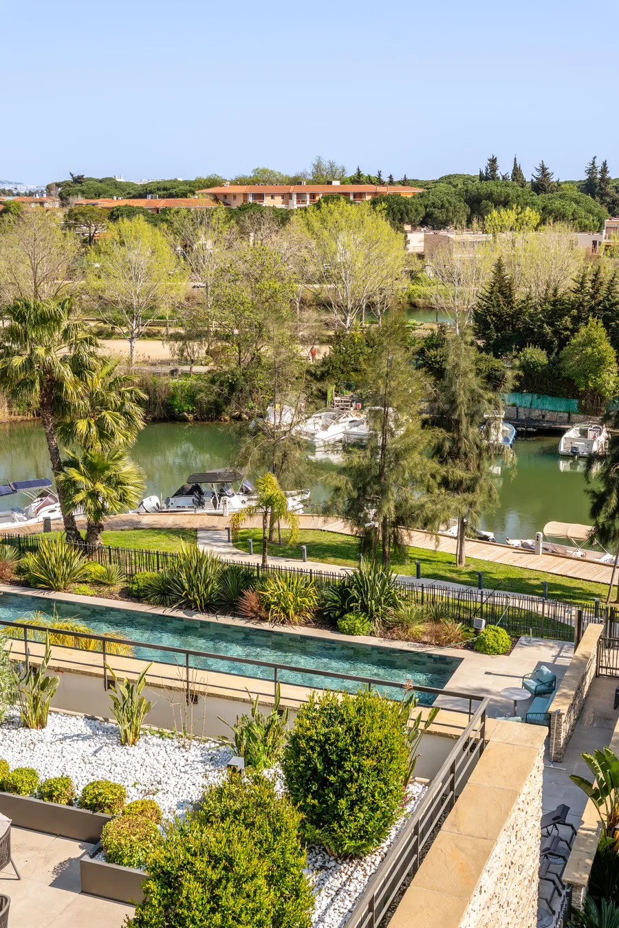 View of a garden with a pool, bordered by trees and plants, overlooking a body of water with moored boats and houses in the background