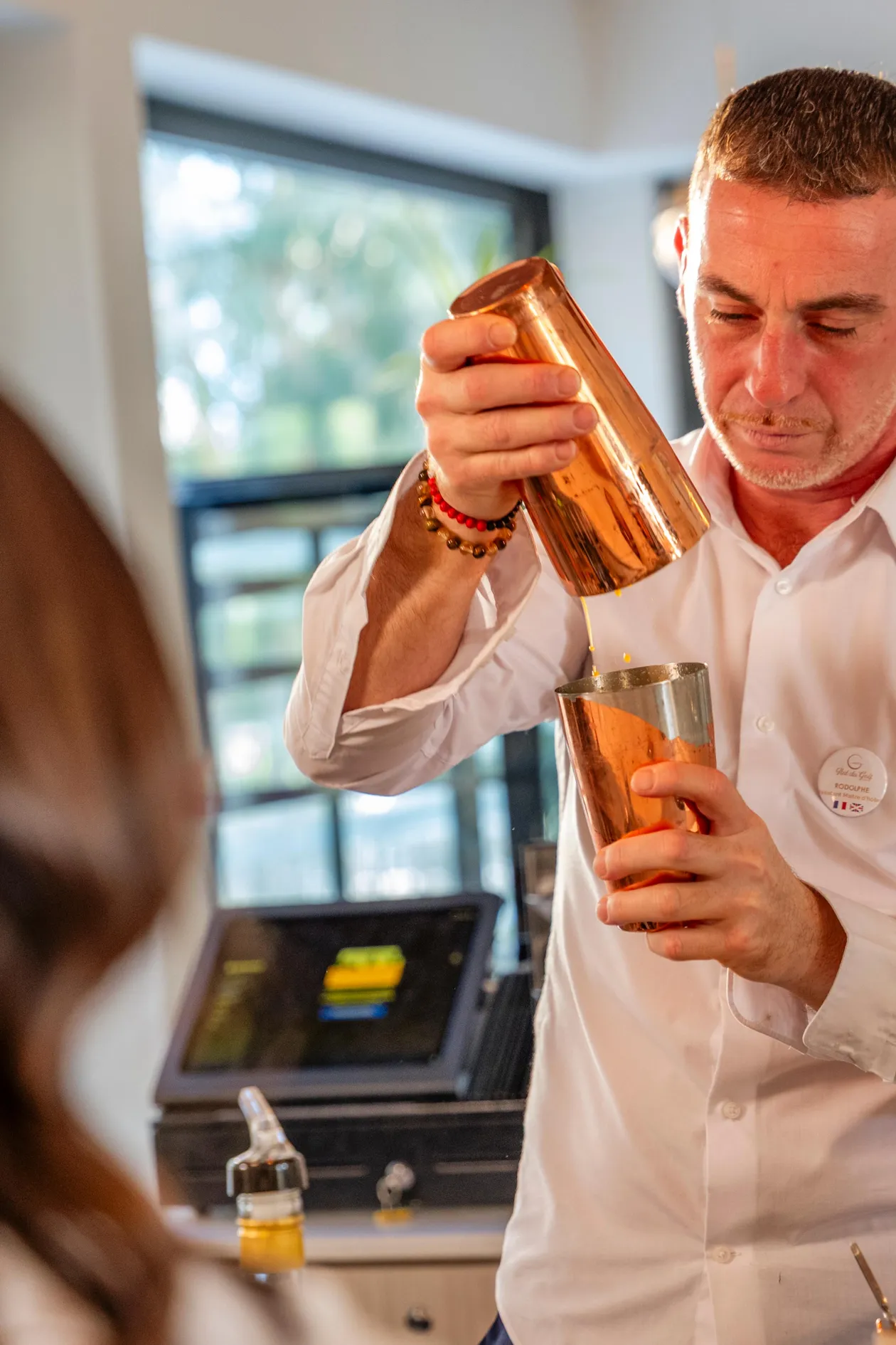 A man in a white shirt prepares a cocktail by pouring a liquid from a metal shaker into a glass.