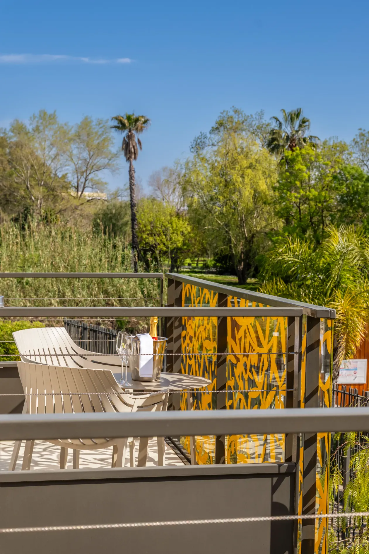 Two deck chairs and a table with glasses and a champagne bucket on a balcony with a metal railing and a yellow décor, surrounded by trees and vegetation.