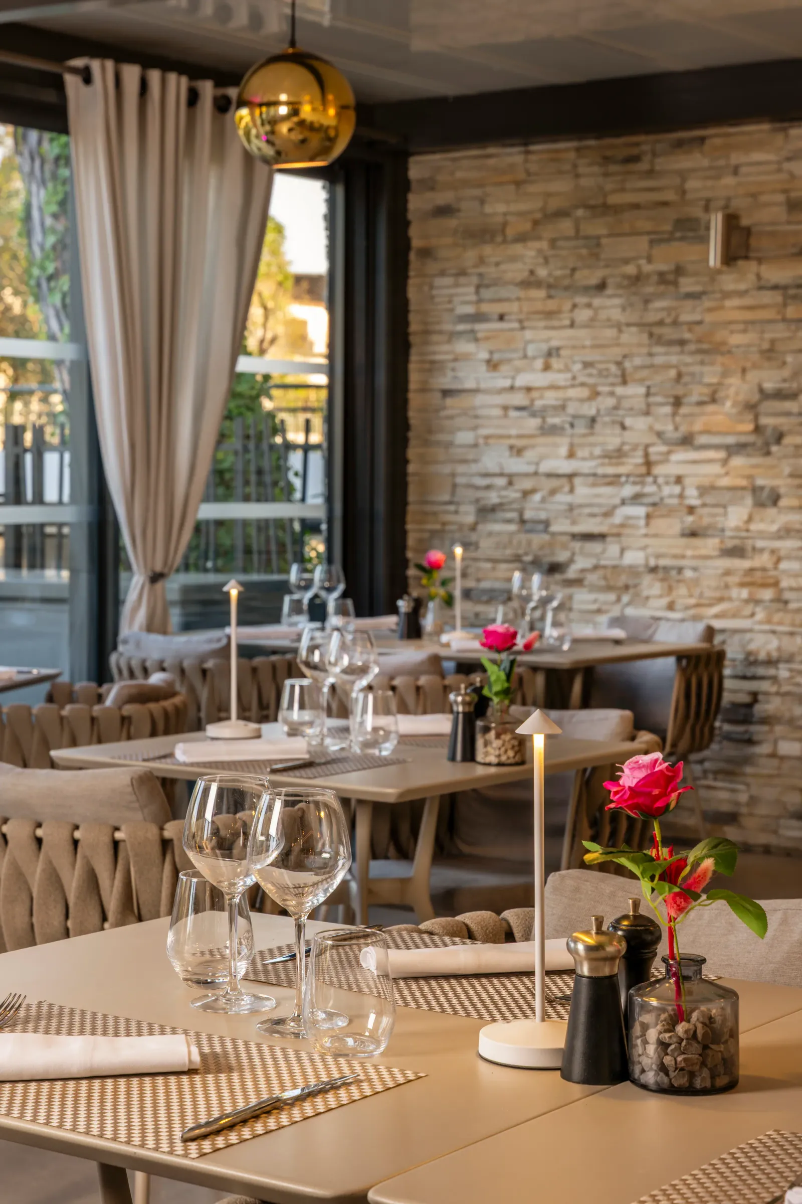 Table set in a restaurant with wine glasses, napkins, flowers, and a salt stone, in front of a stone wall and a window with curtains.