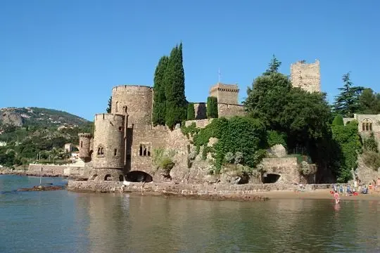 Medieval stone castle located by a body of water, surrounded by trees and vegetation, with a beach and people in the background.