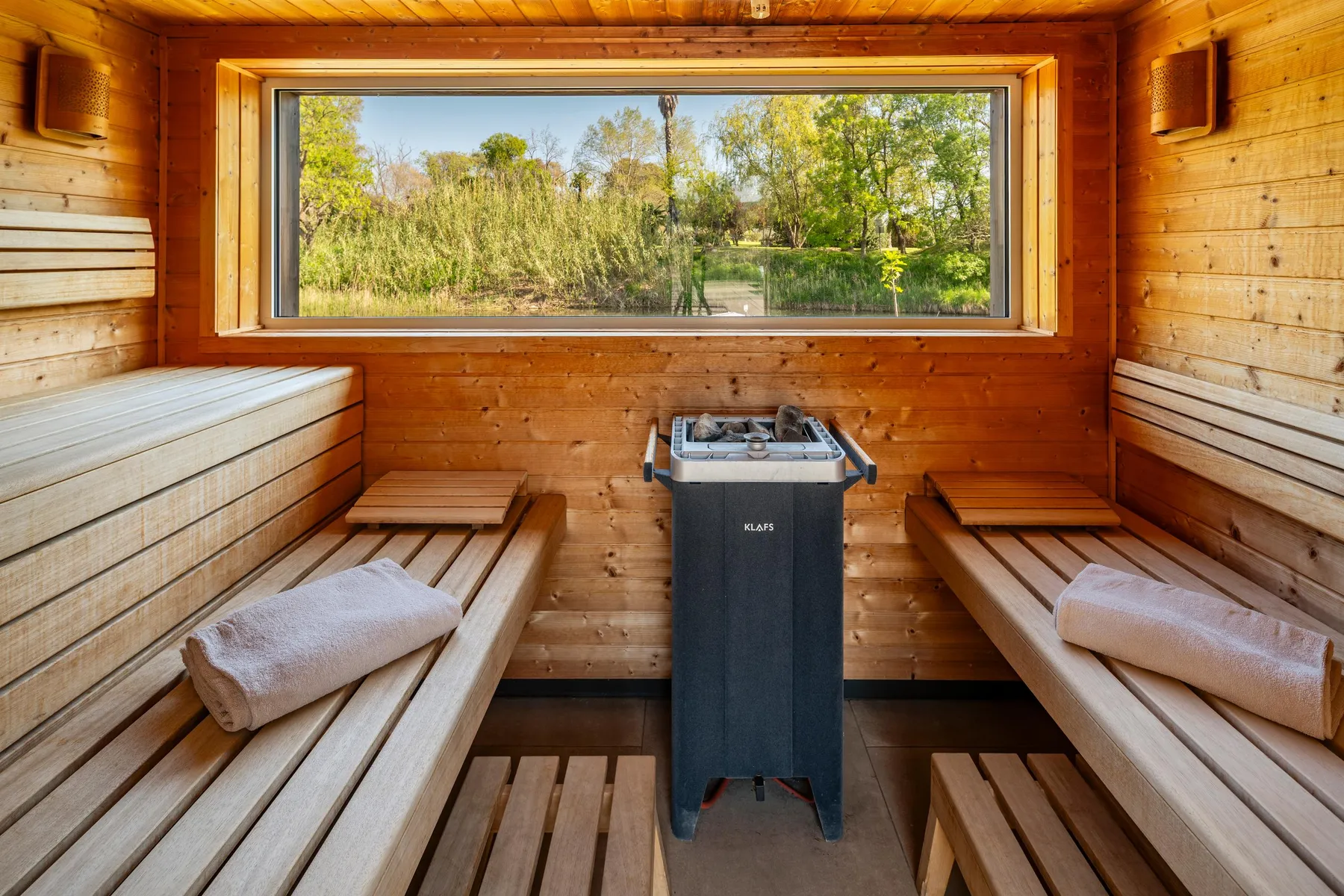 Wooden sauna with benches, towels, and a heating stove, offering an outdoor view through a window.