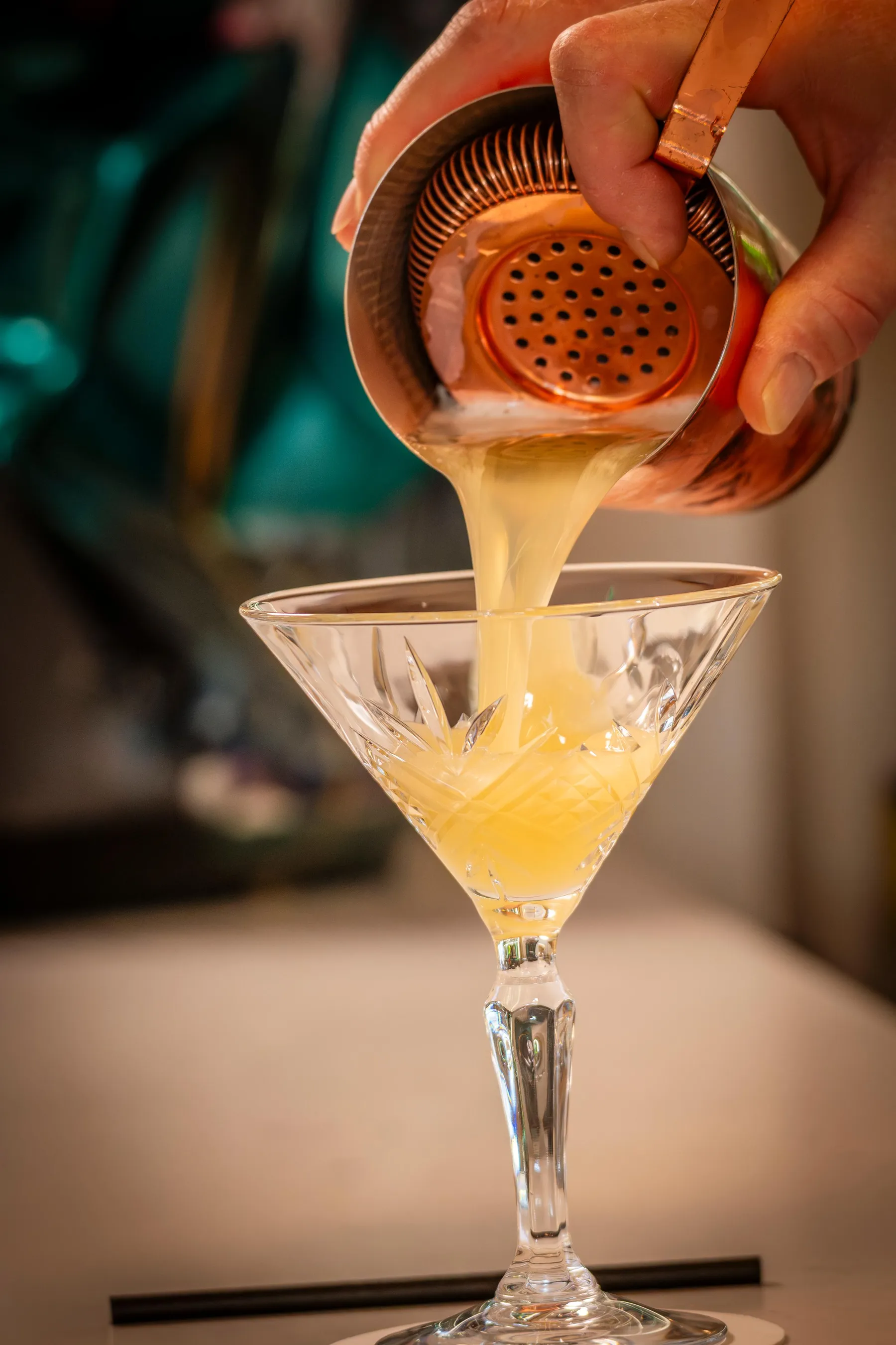 A yellow liquid is poured into a martini glass using a cocktail strainer.