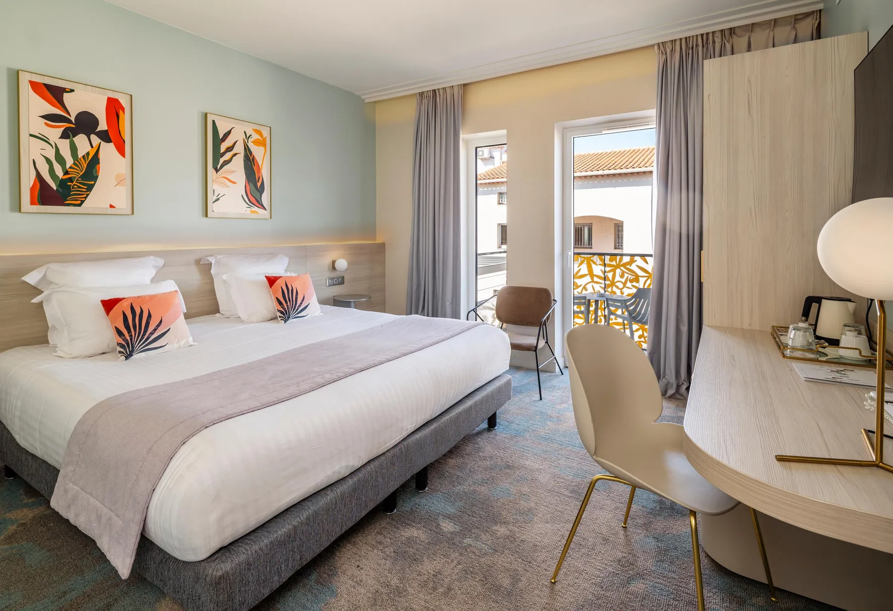 Hotel room with a double bed, a bedside table, a chair, and a French door leading to a balcony with a view of rooftops.