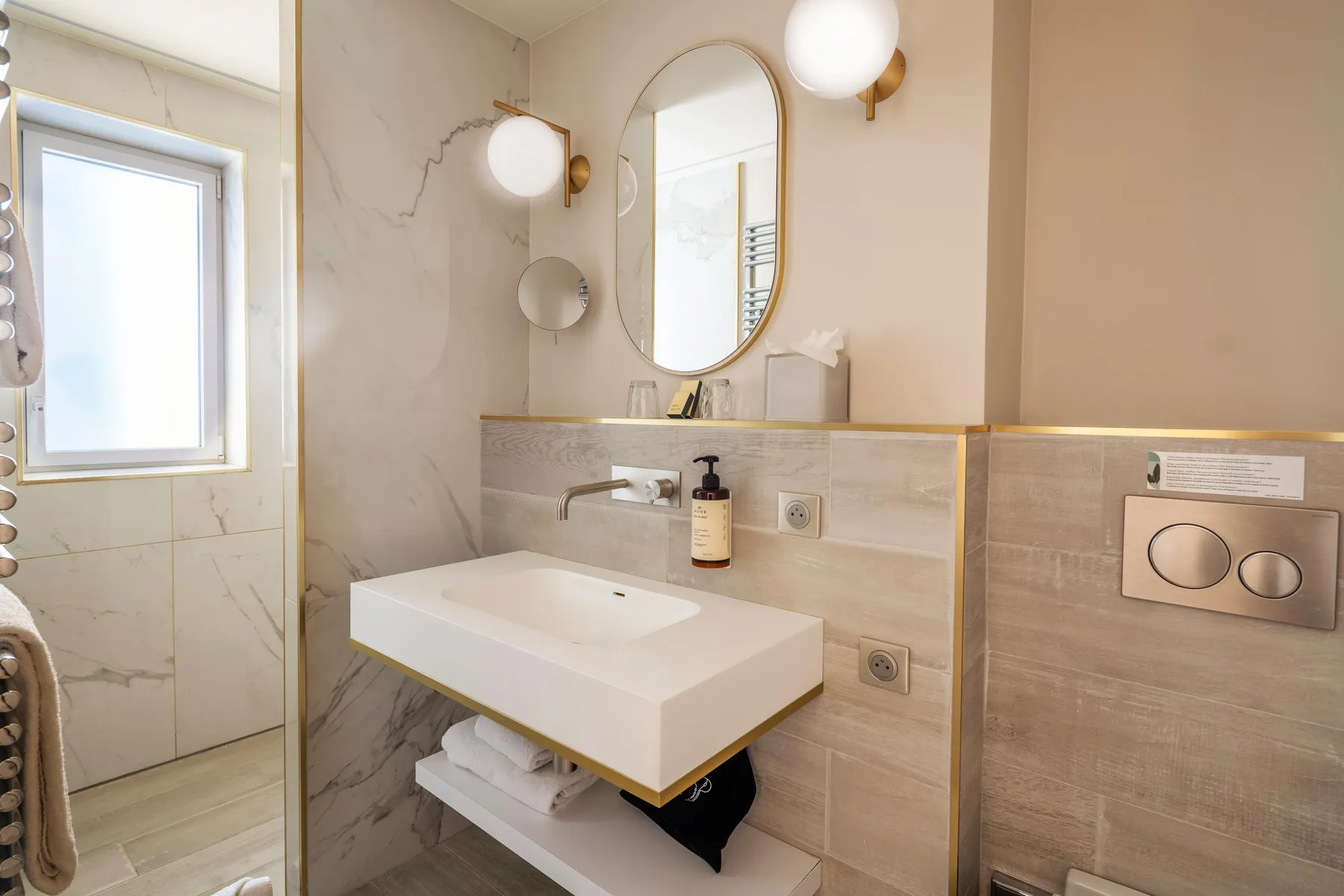 A white sink with faucetry, oval and round mirrors, spherical lights, a window, and shelves in a bathroom with beige and marble tiled walls.