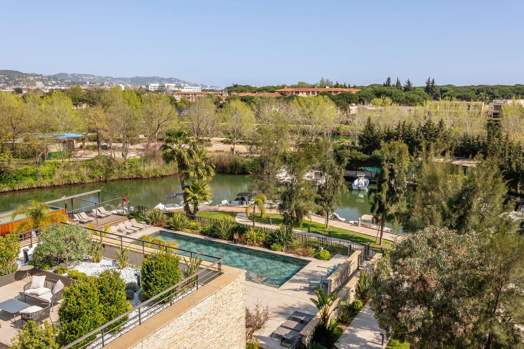 Aerial view of a garden with a pool, bordered by trees and a body of water where boats are moored.
