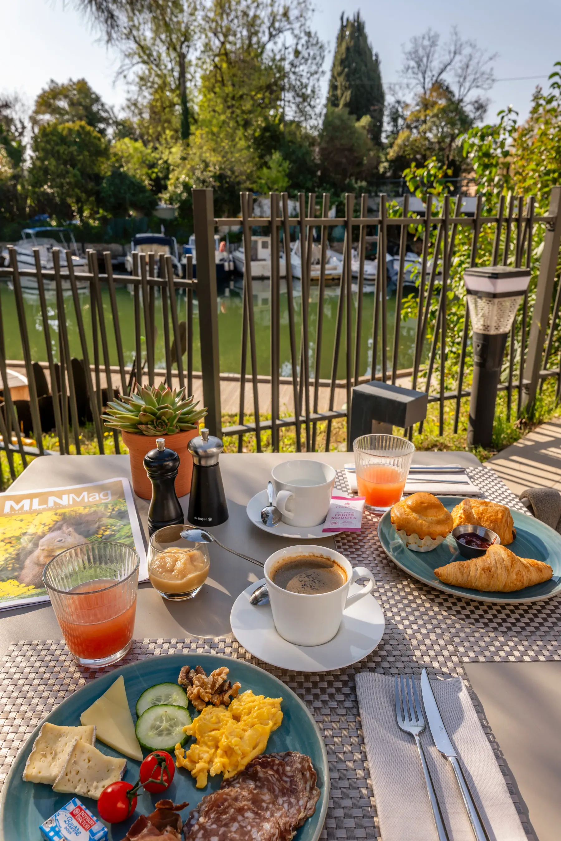 Table de petit-déjeuner en extérieur avec café, jus d'orange, croissants, œufs brouillés, viande, fromage, pain, tomates et concombres, vue sur un jardin et des arbres