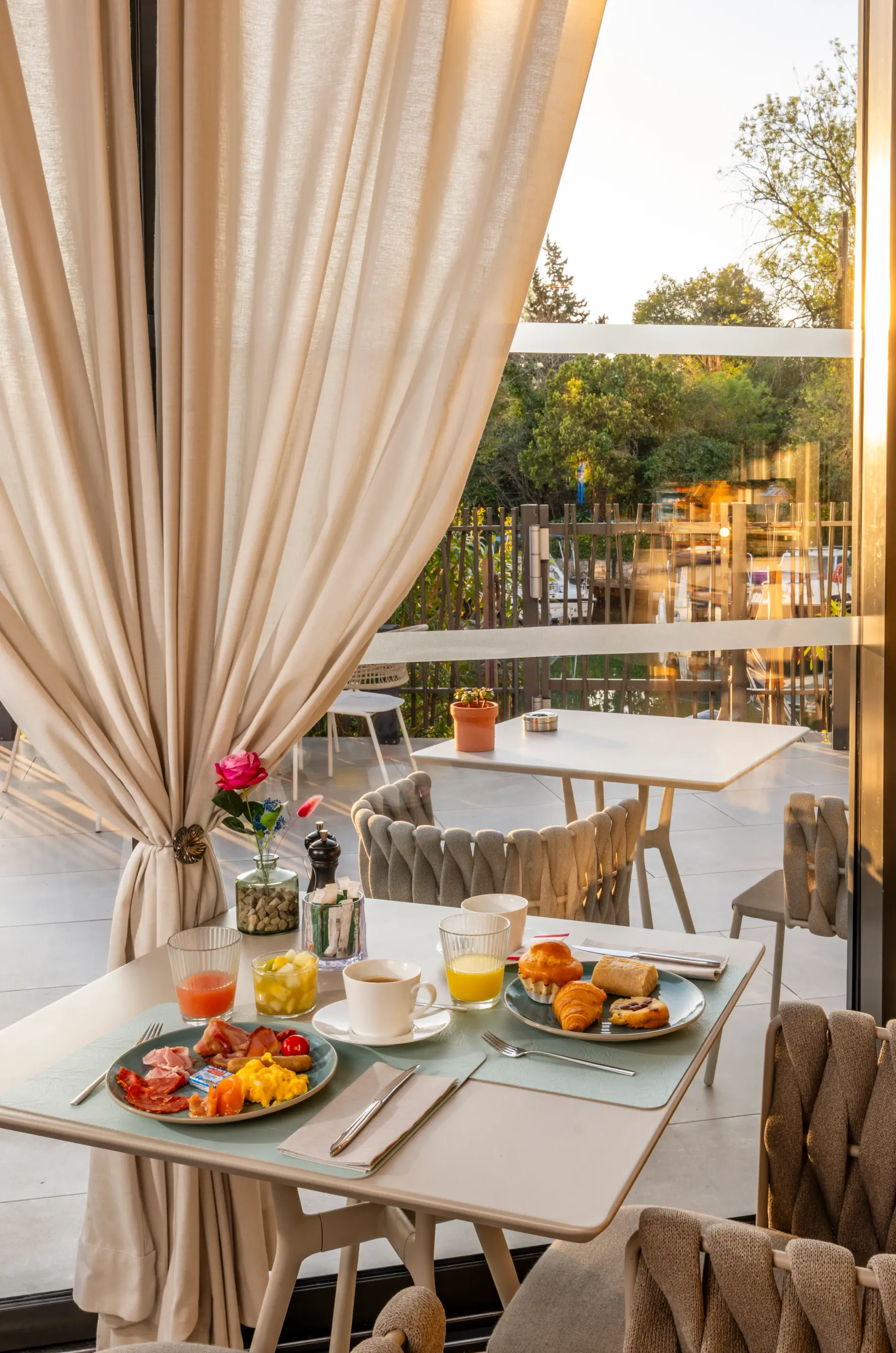 Table dressée pour le petit-déjeuner avec des plats de charcuterie, des croissants, des fruits, des boissons et une rose dans un vase, vue depuis l'intérieur d'un restaurant avec des rideaux et une terrasse extérieure visible à travers une baie vitrée