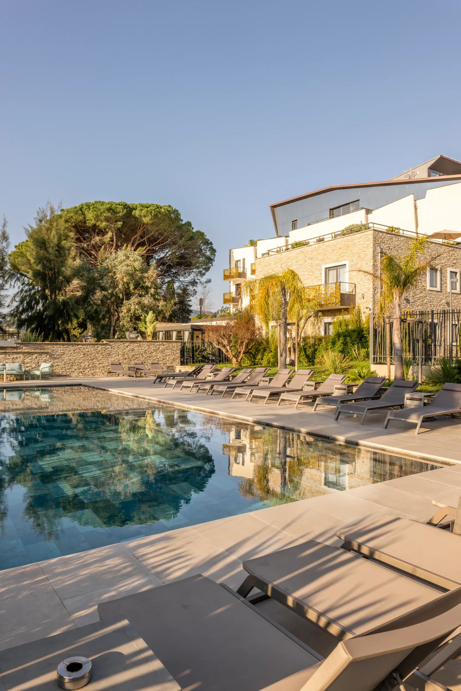 Pool surrounded by sun loungers with a stone building and trees in the background