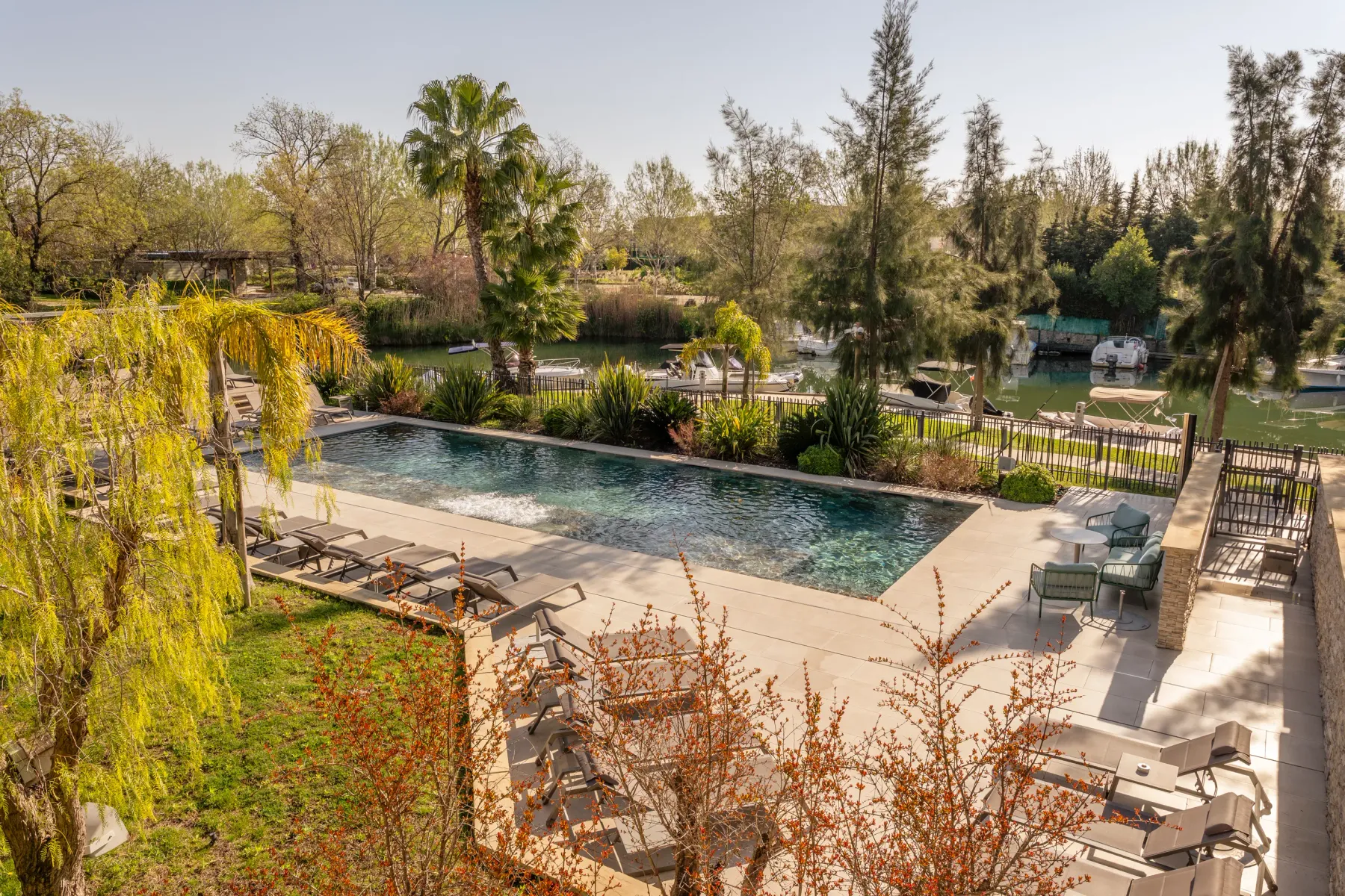 Pool surrounded by sun loungers and tables, with a body of water and boats in the background, in a tree-lined setting.