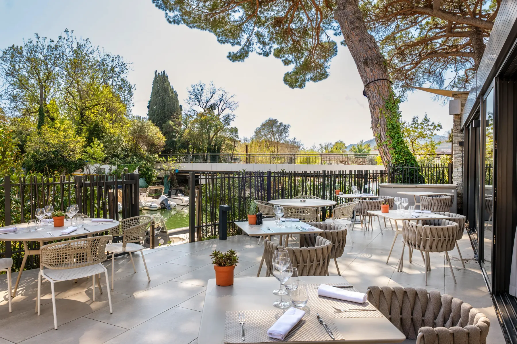 Restaurant terrace with tables and chairs arranged under a pergola, surrounded by trees and greenery.