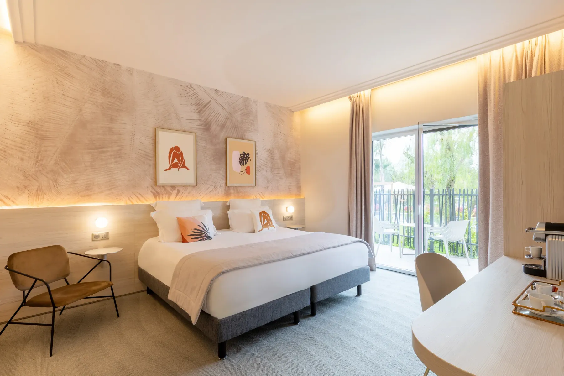 Modern hotel room with a double bed, a table with two chairs, an armchair, a sliding door leading to a balcony, and artwork on the wall.
