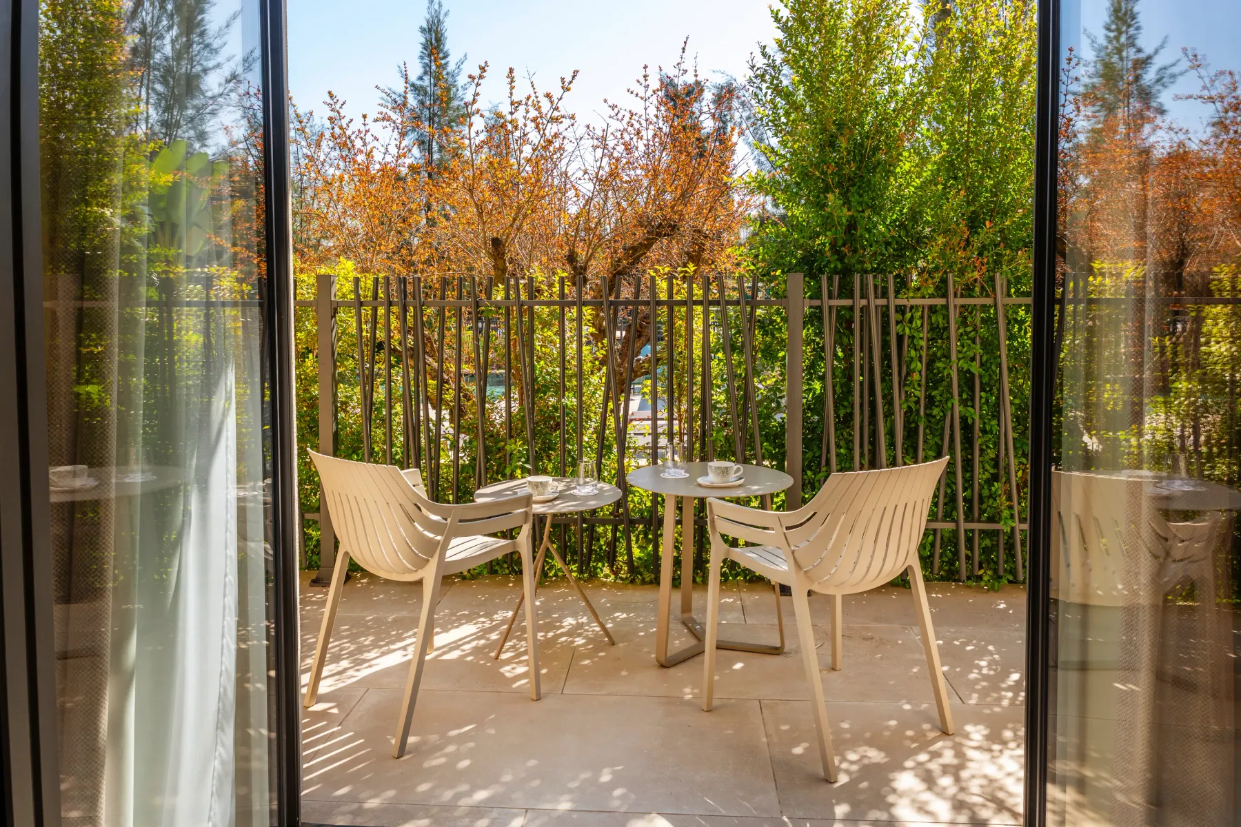 Two chairs and a round table on a stone terrace, surrounded by dense vegetation and a metal fence, seen through a glass door.
