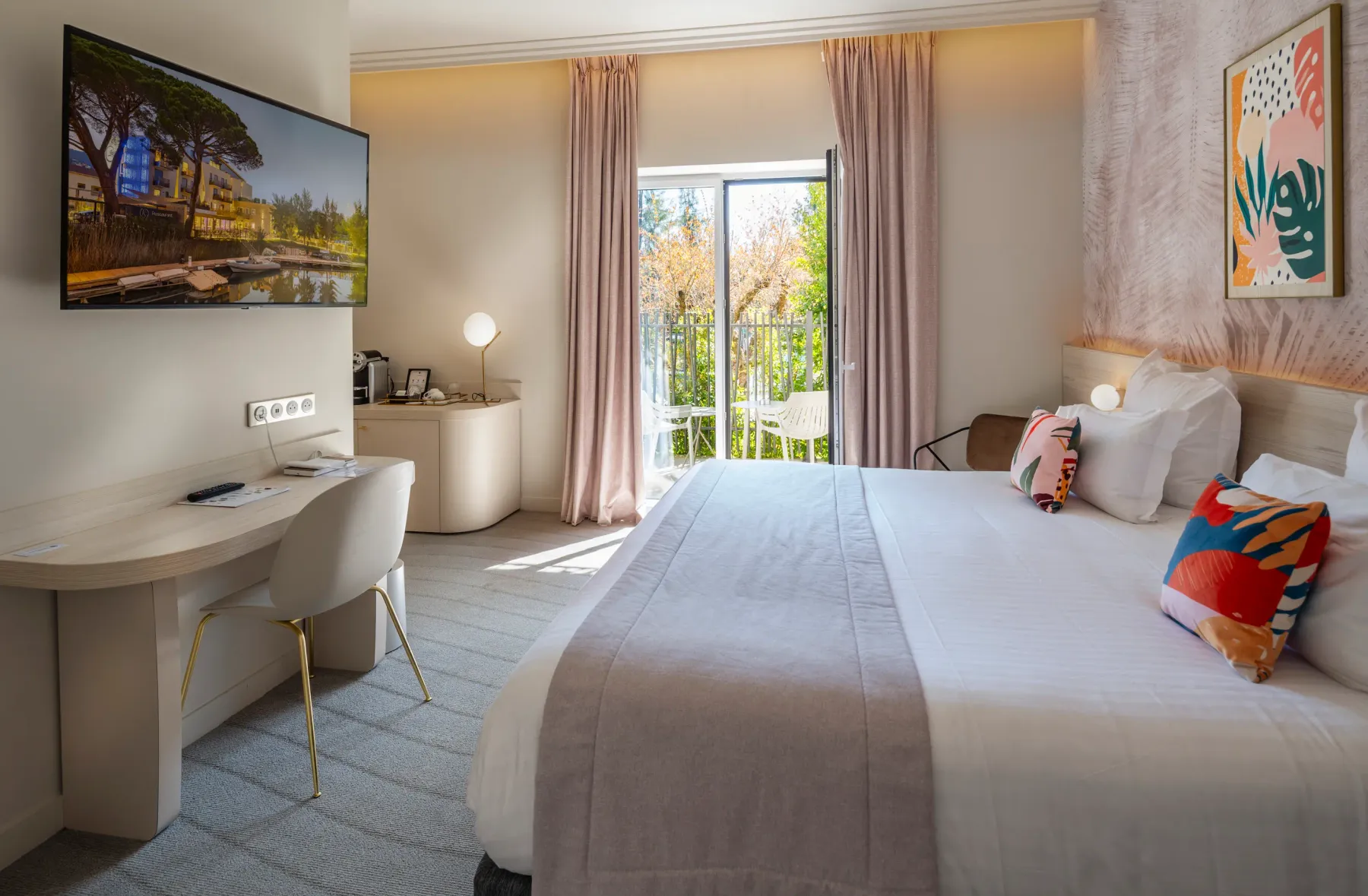 Hotel room with a bed, a desk, a television, a French window leading to a balcony, and artwork on the walls.