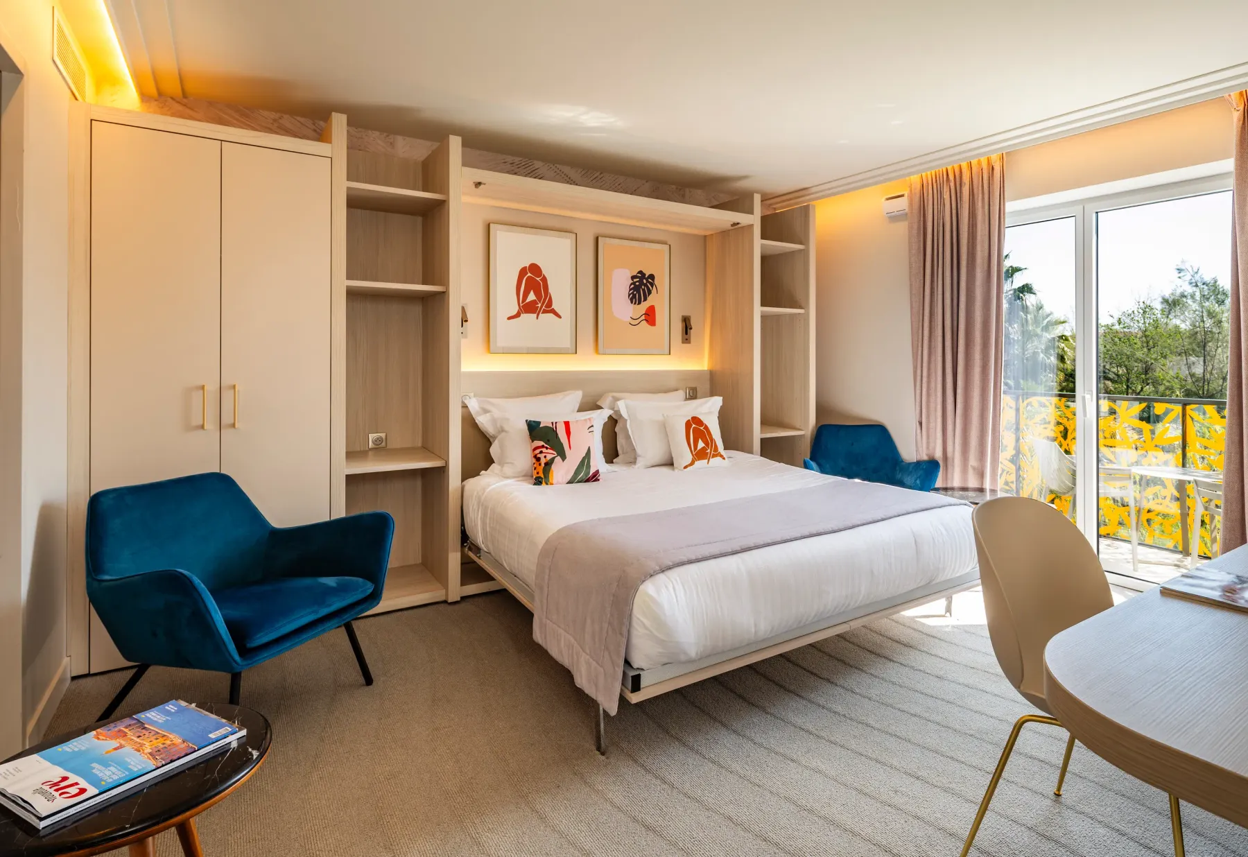 Hotel room with a double bed, two armchairs, a round table, and a view of a balcony with plants.