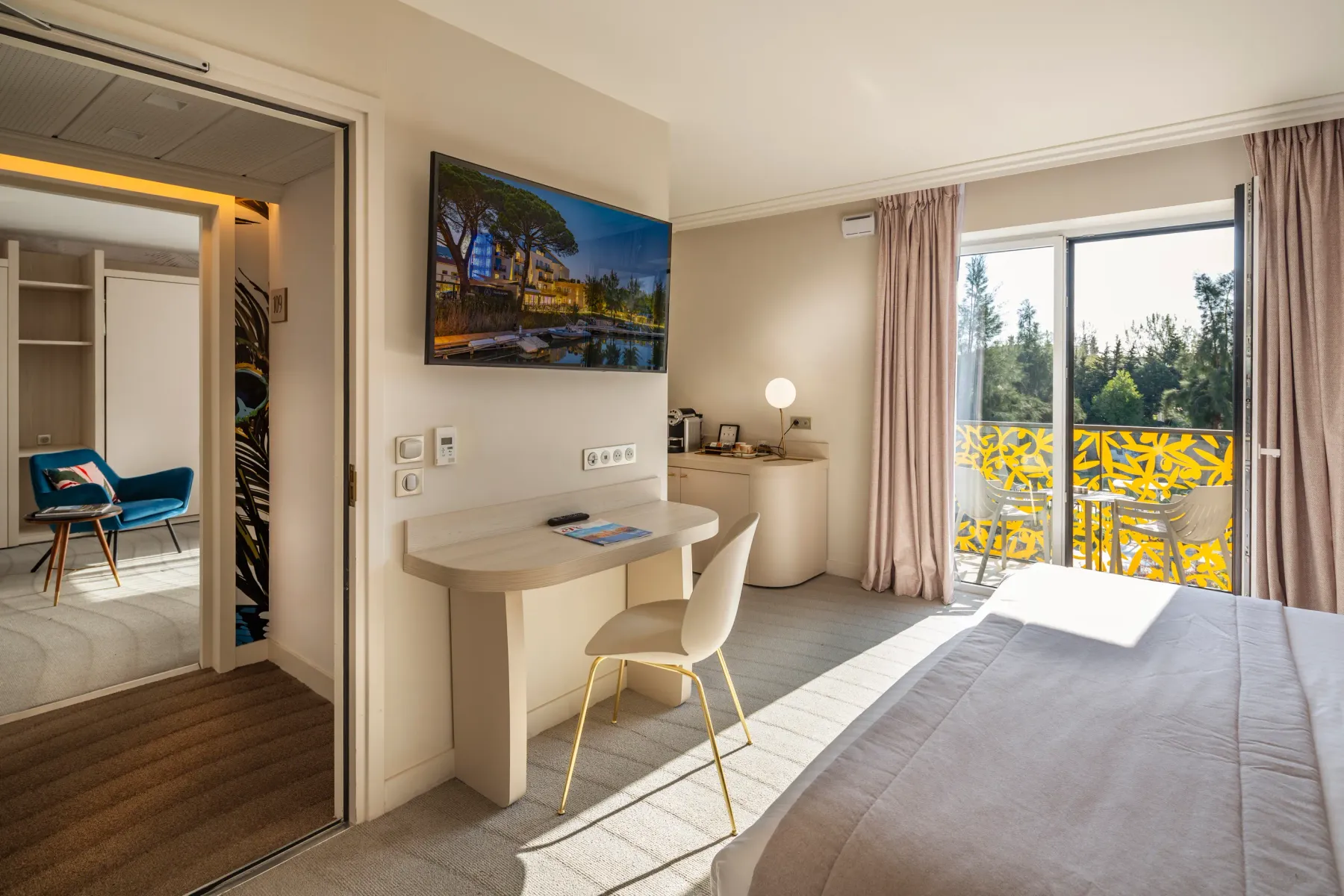 Modern hotel room with a bed, a desk, a television, a seating area, and a sliding door leading to a balcony with a view of nature.