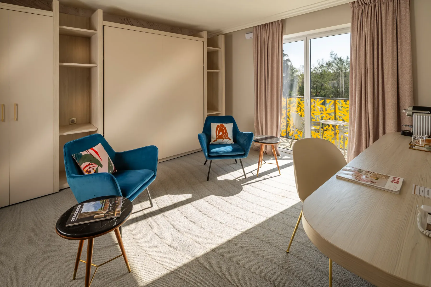 Room with two blue armchairs, a round table, a desk, shelves, and a French window opening onto a balcony with a view of trees.