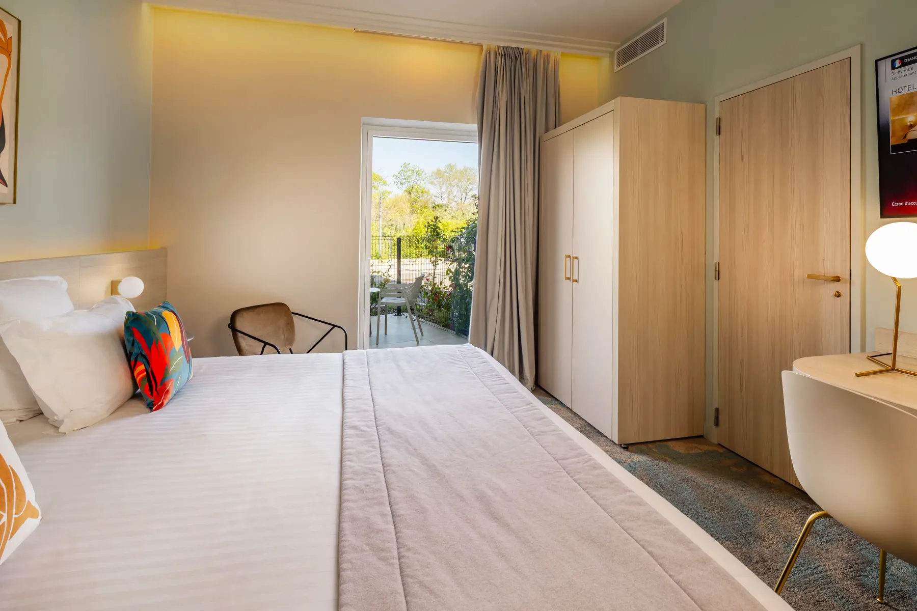 Hotel room with a double bed, a French window leading to a balcony, wardrobes, and a bedside table with a lamp.