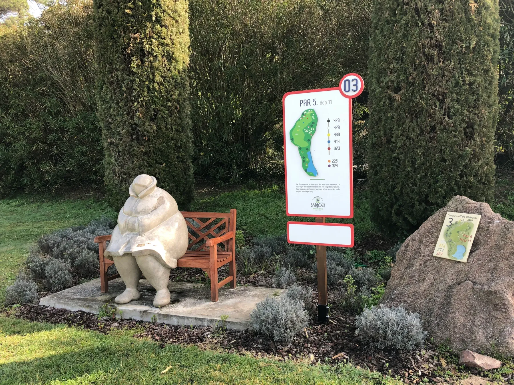 Statue d'une personne assise sur un banc devant un panneau de golf indiquant le trou numéro 3, par 5, avec un diagramme du parcours